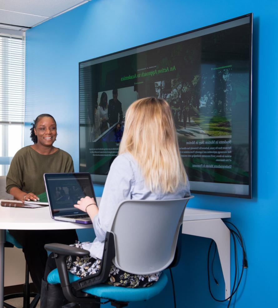 Two women seating at a desk with a laptop between them.